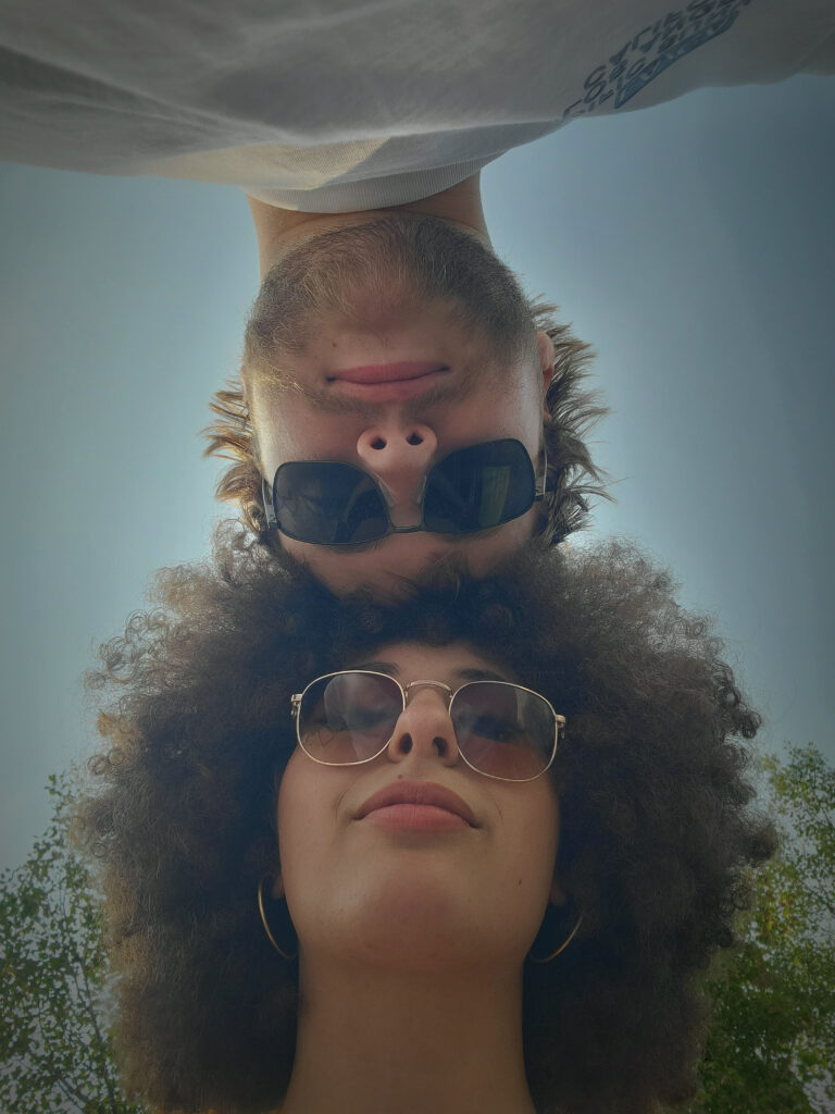 An image of a woman with an afro and blonde man, both wearing sunglasses on a sunny day, looking down at the ground/camera as the top of their heads touch.