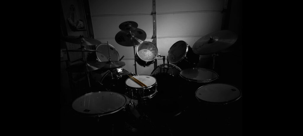 a black and white image of a large double bass drum kit with multiple symbols. Resting on the snare drum are two tan, thin drumsticks.