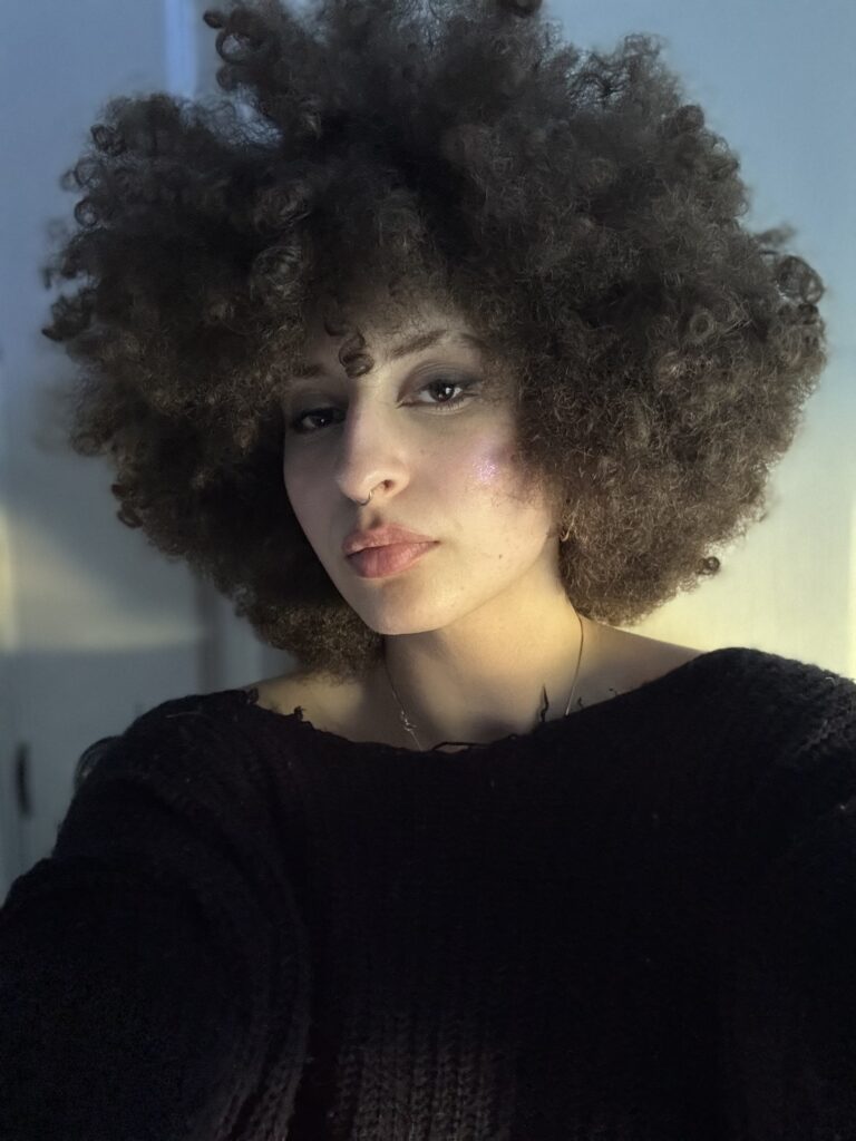 A woman with curly hair is taking a selfie facing the camera at an angle. wearing a blue sweater in a white room